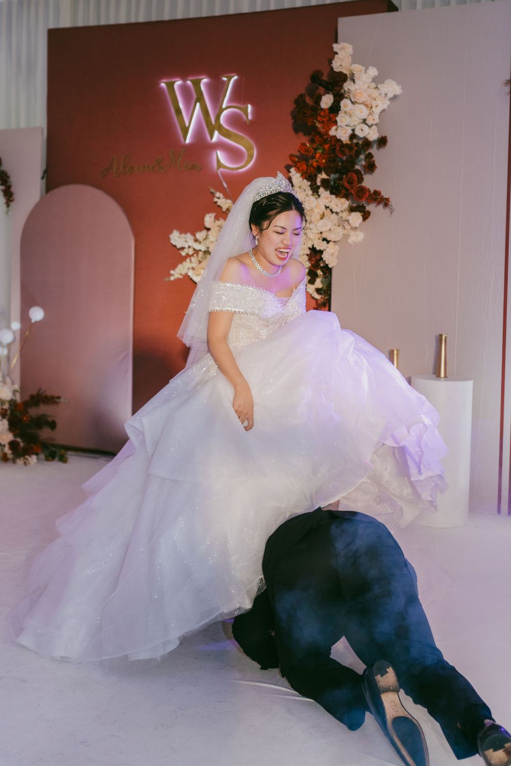 Playful wedding photo shoot moment with bride in white gown against terracotta-colored backdrop.