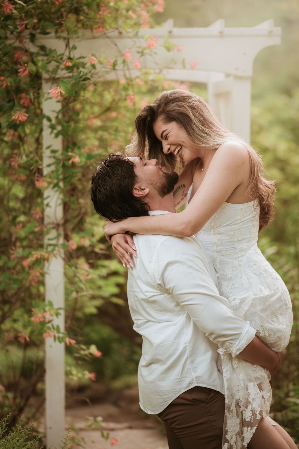 A joyful couple embraces in a romantic garden, the man lifting the laughing woman near a floral trellis.