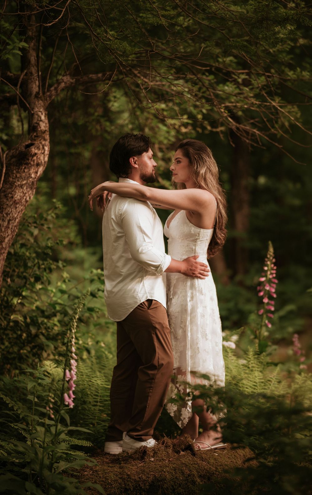 A couple embraces romantically in a lush forest setting, surrounded by ferns and foxglove flowers.