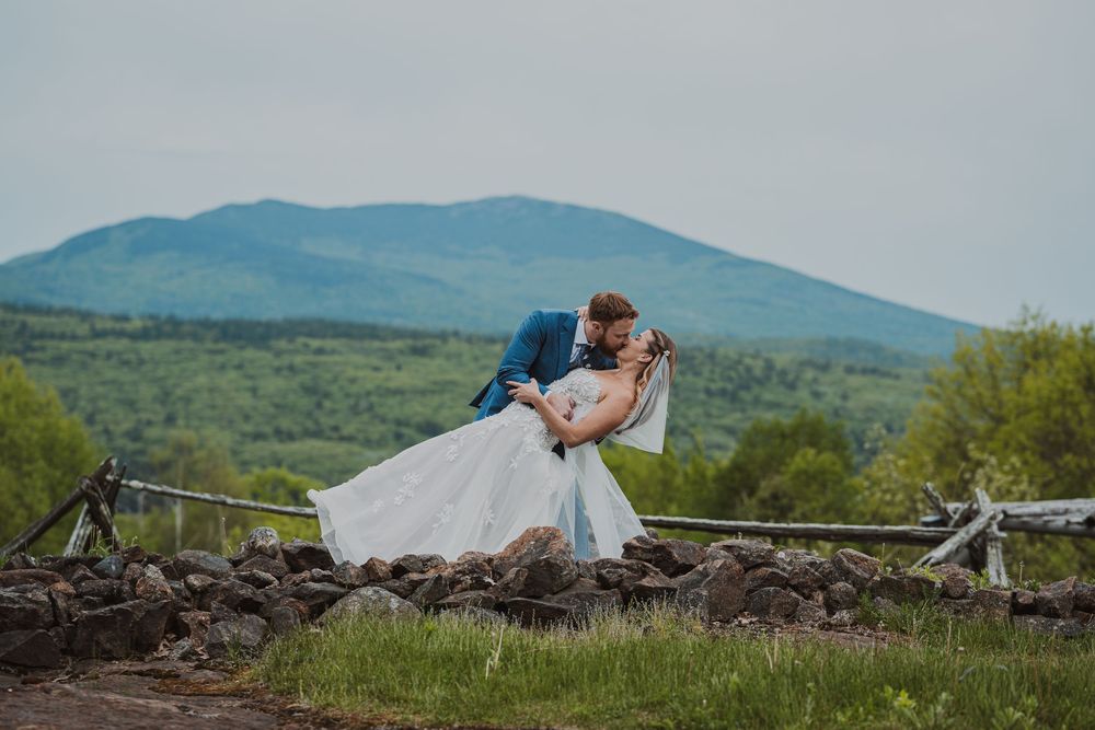 Bride and groom share a romantic dip kiss with lush green mountains and stone fence backdrop on their wedding day.