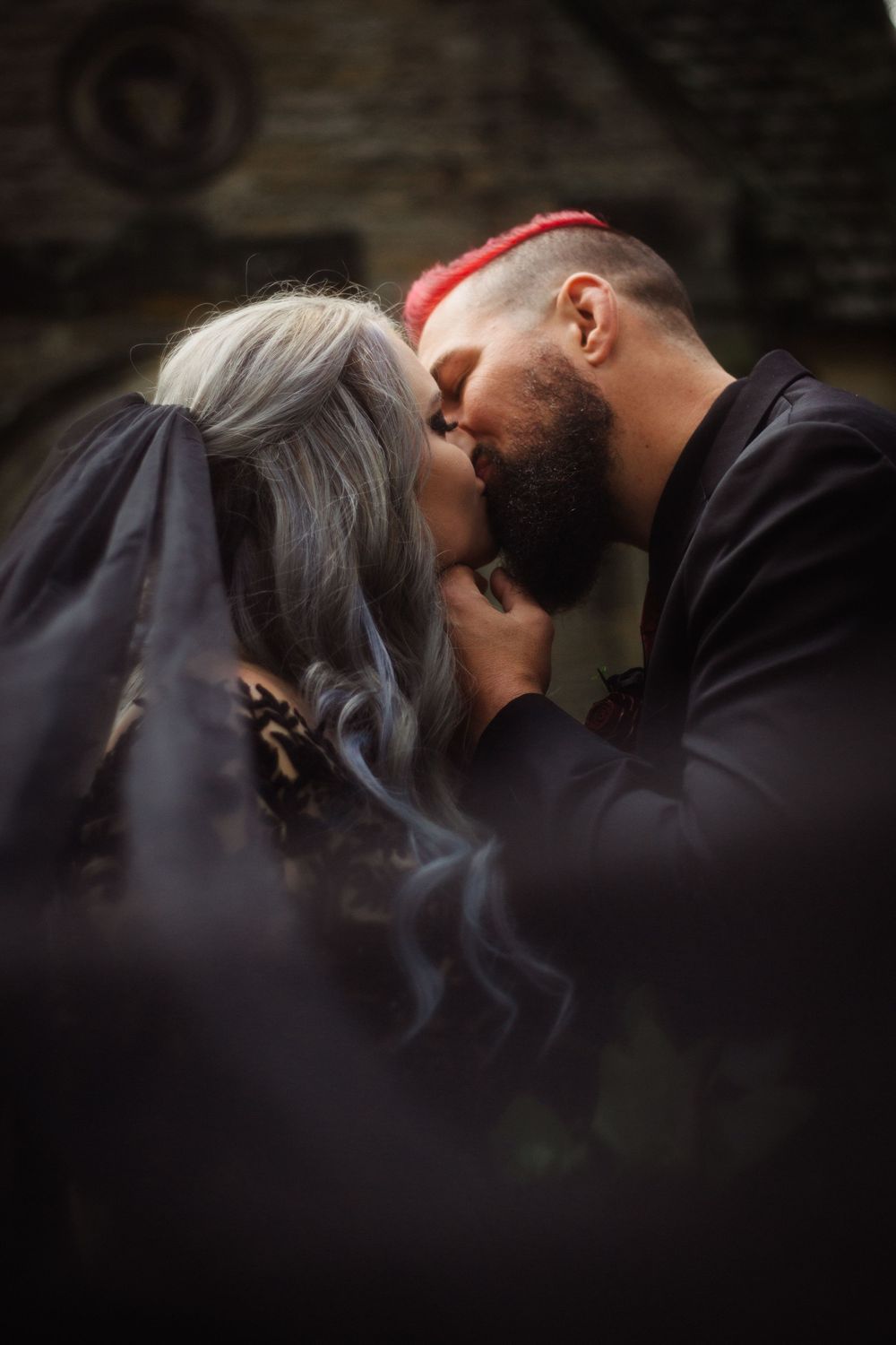 Couple in dark wedding attire sharing a kiss against a stone backdrop.