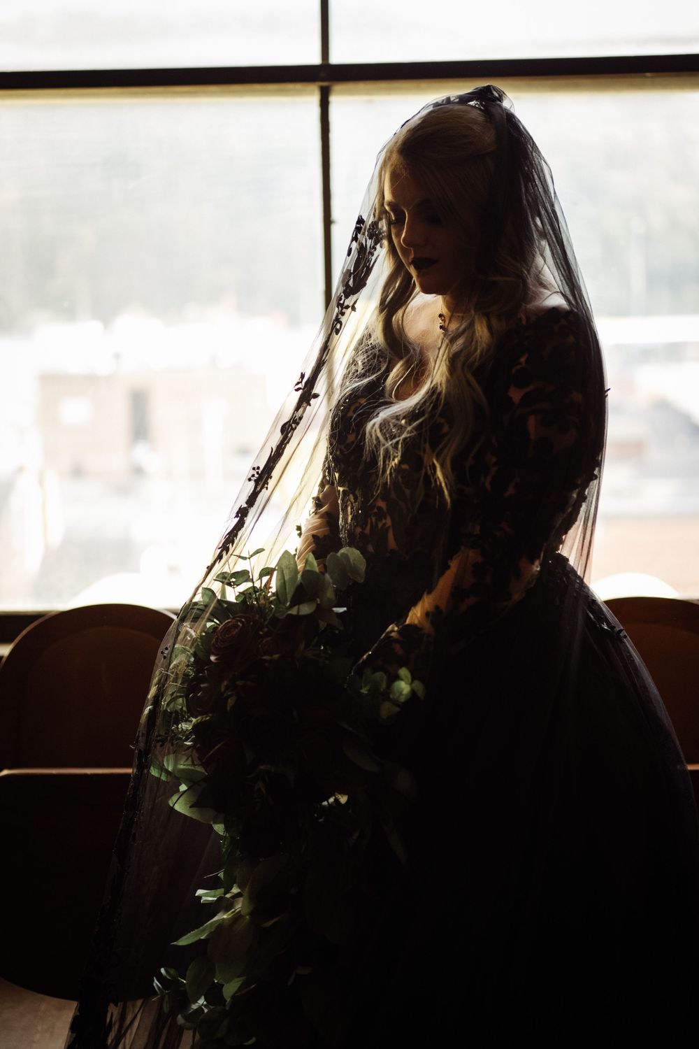 Silhouette of bride in black gown and veil holding floral bouquet by window.