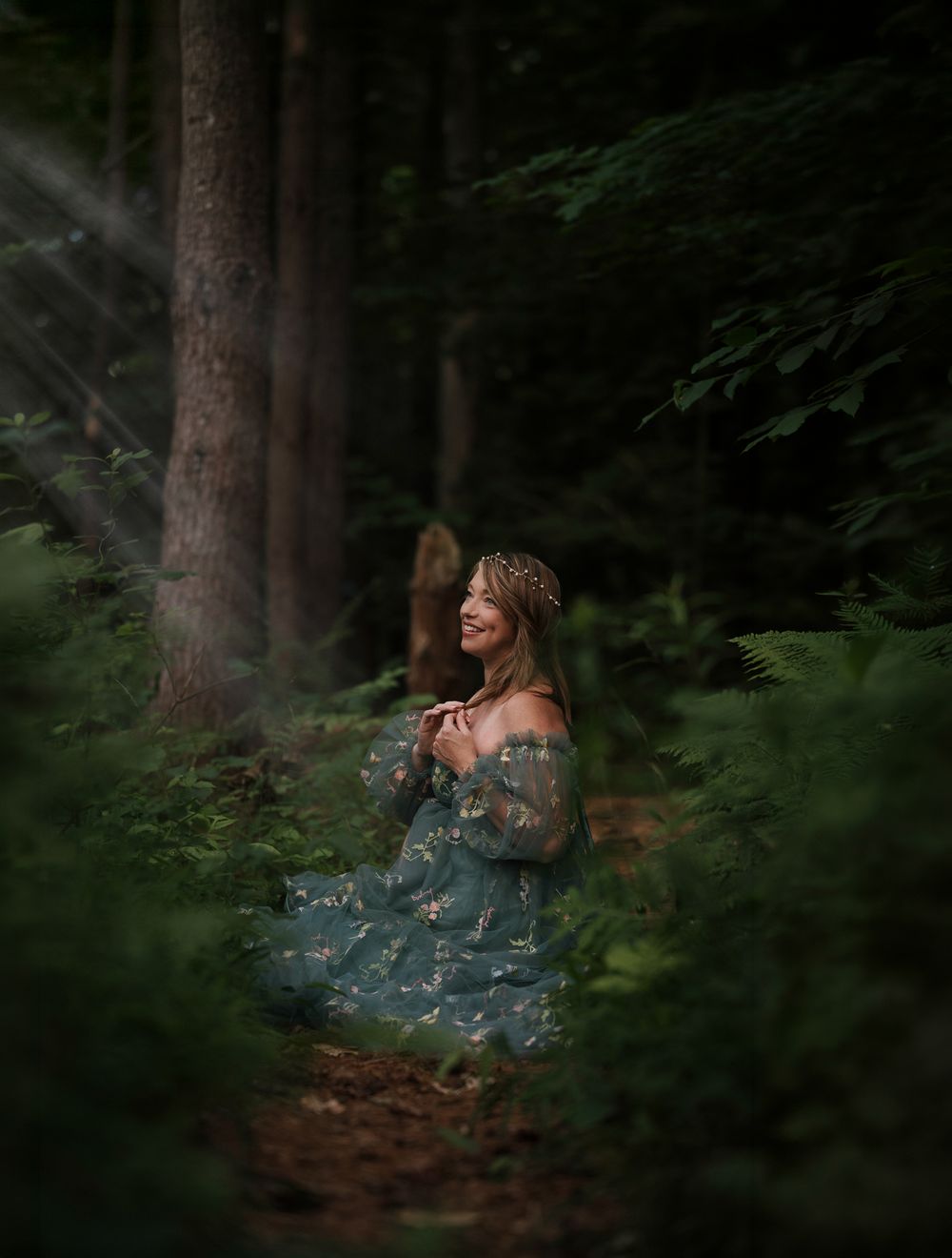 A woman in a teal floral gown and flower crown smiles in a sunlit forest clearing.