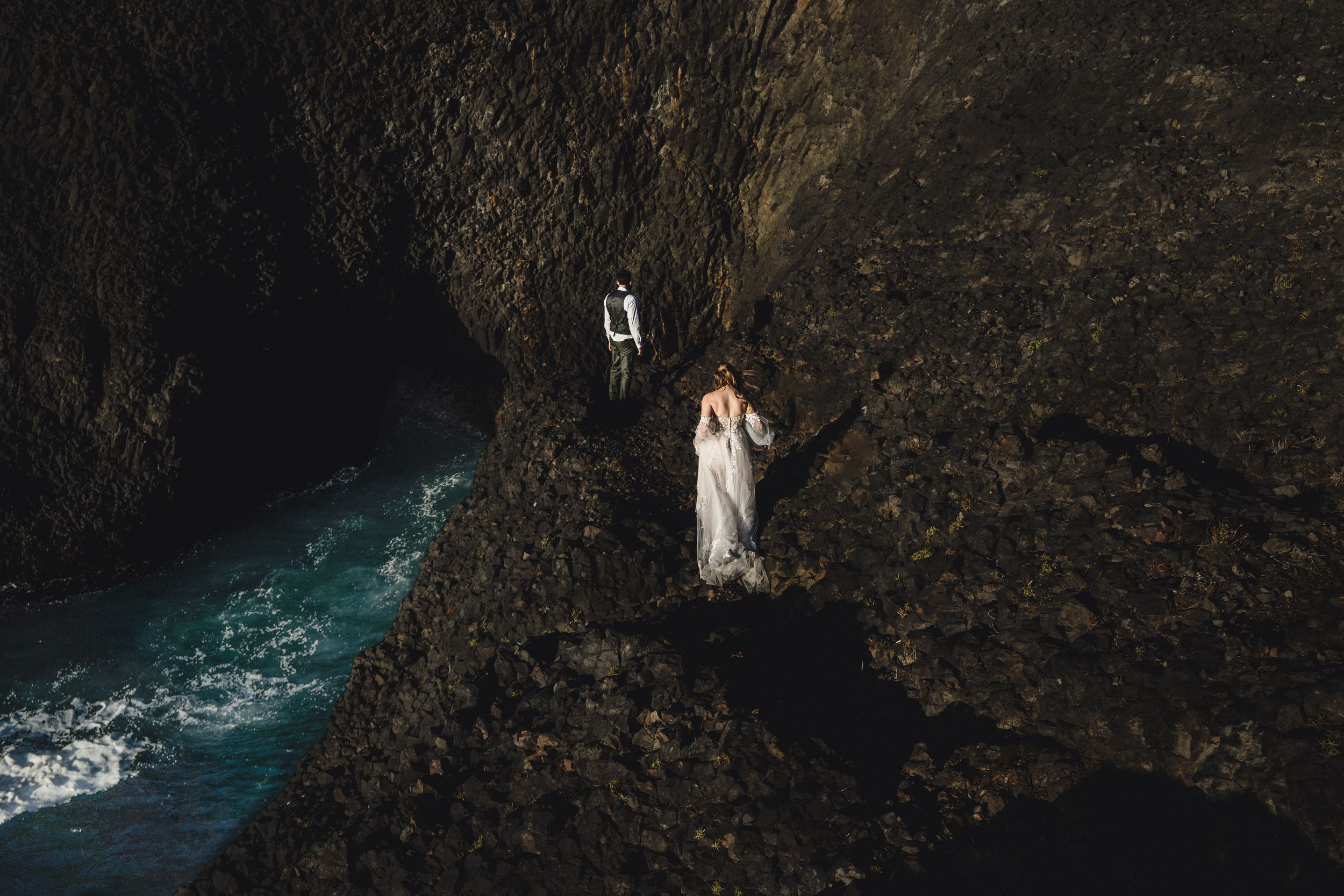Bride and groom in wedding attire stand on dramatic rocky coastal cliffs above turquoise ocean waves.