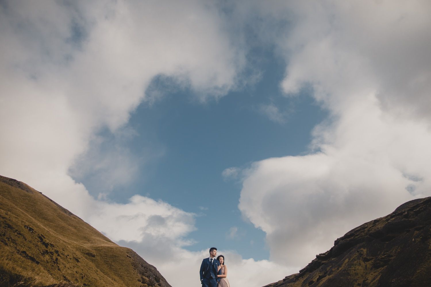 Couple embracing between rolling green hills under dramatic cloudy blue sky in scenic mountain landscape.