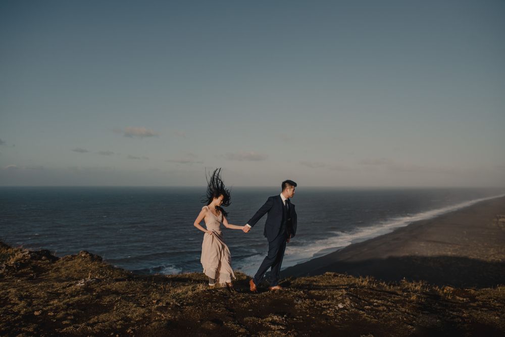 Couple holding hands on a coastal cliff at sunset with ocean waves and black sand beach in the background.