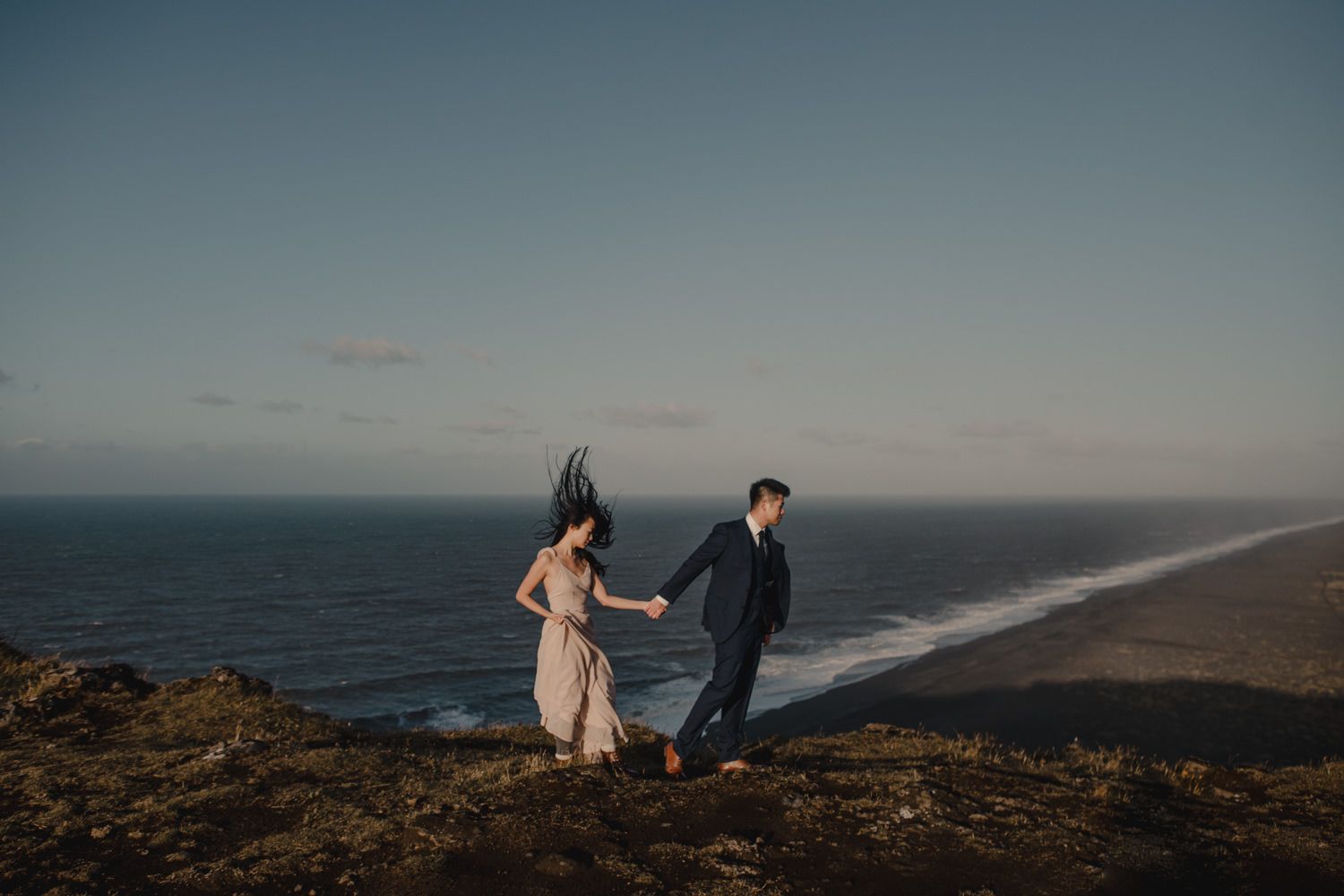 Couple holding hands on a coastal cliff at sunset with ocean waves and black sand beach in the background.