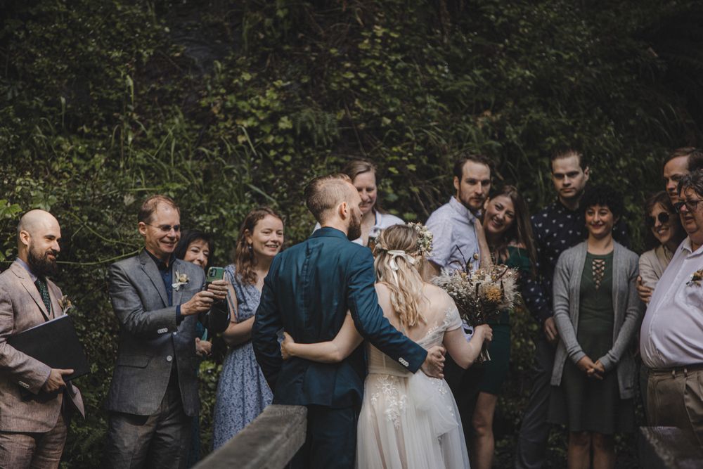 Bride and groom sharing first kiss surrounded by guests at an outdoor wedding ceremony.