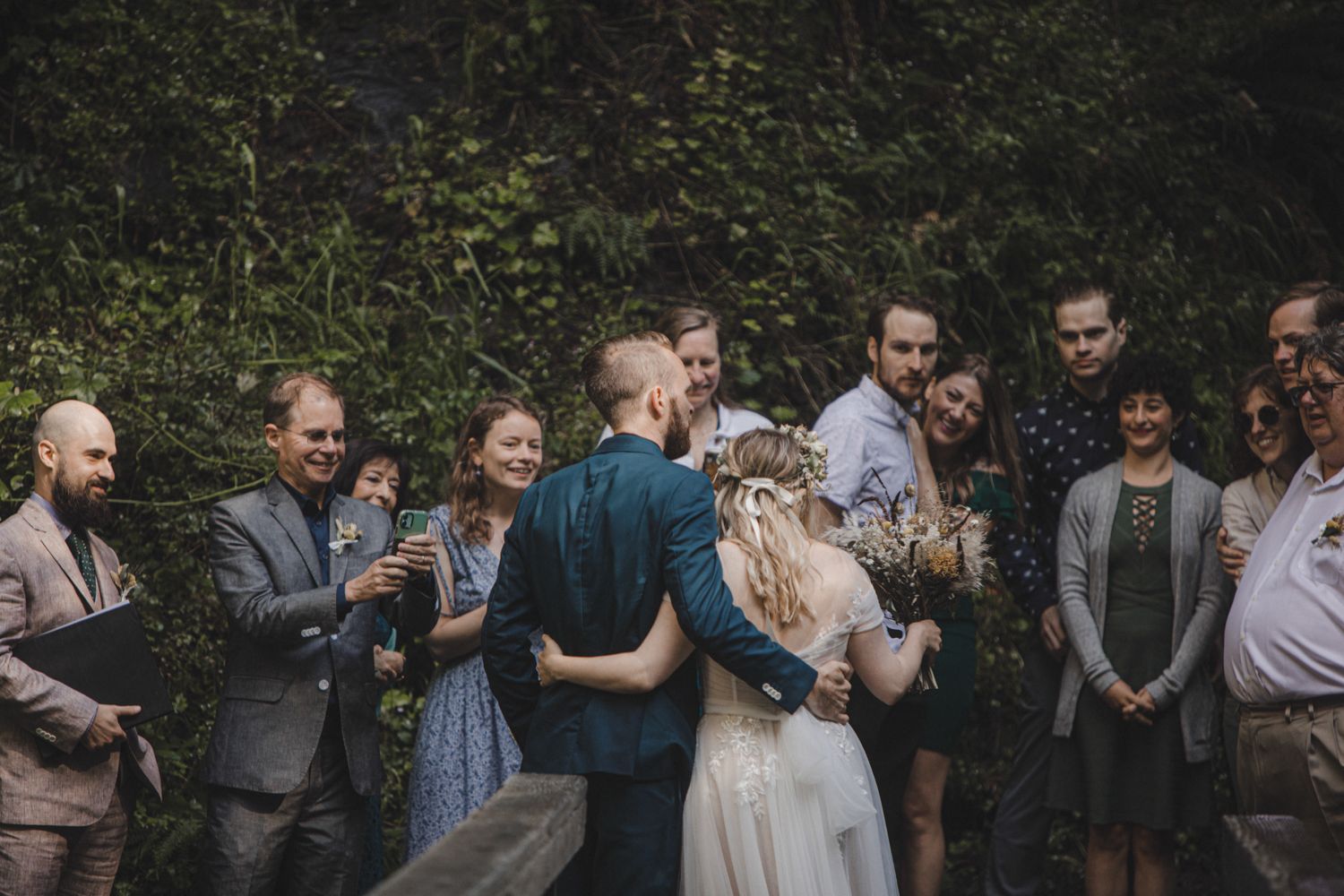 Bride and groom sharing first kiss surrounded by guests at an outdoor wedding ceremony.