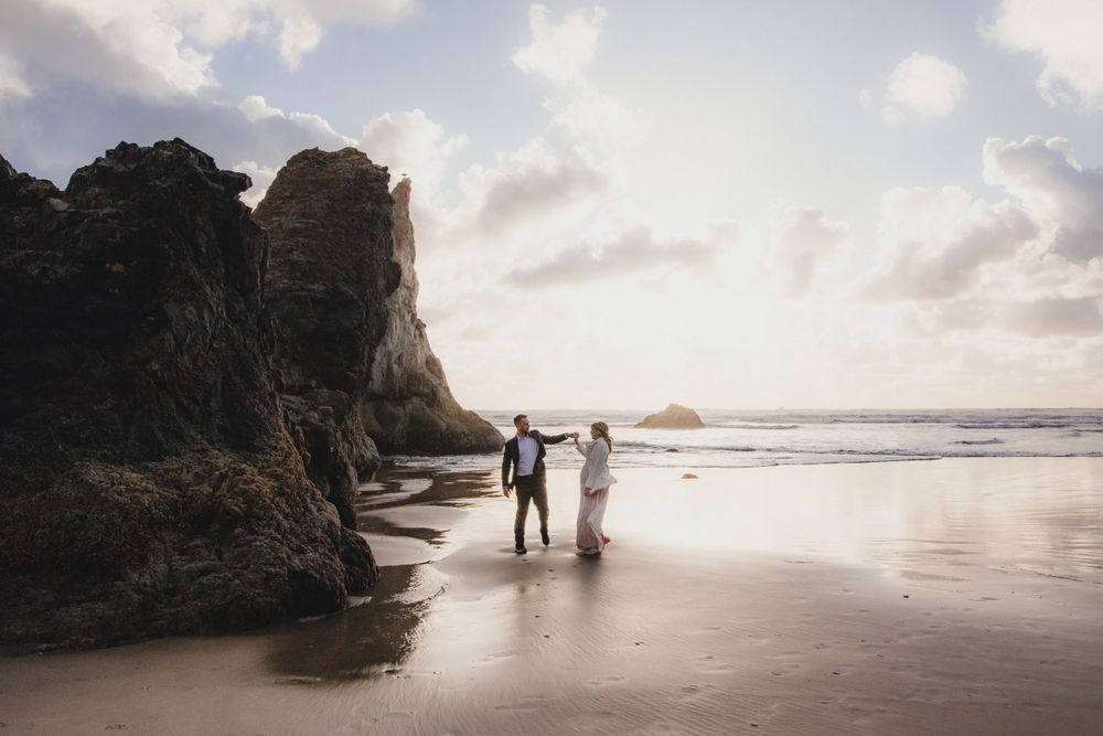 A couple walks along a scenic beach at sunset with towering cliffs in the background.