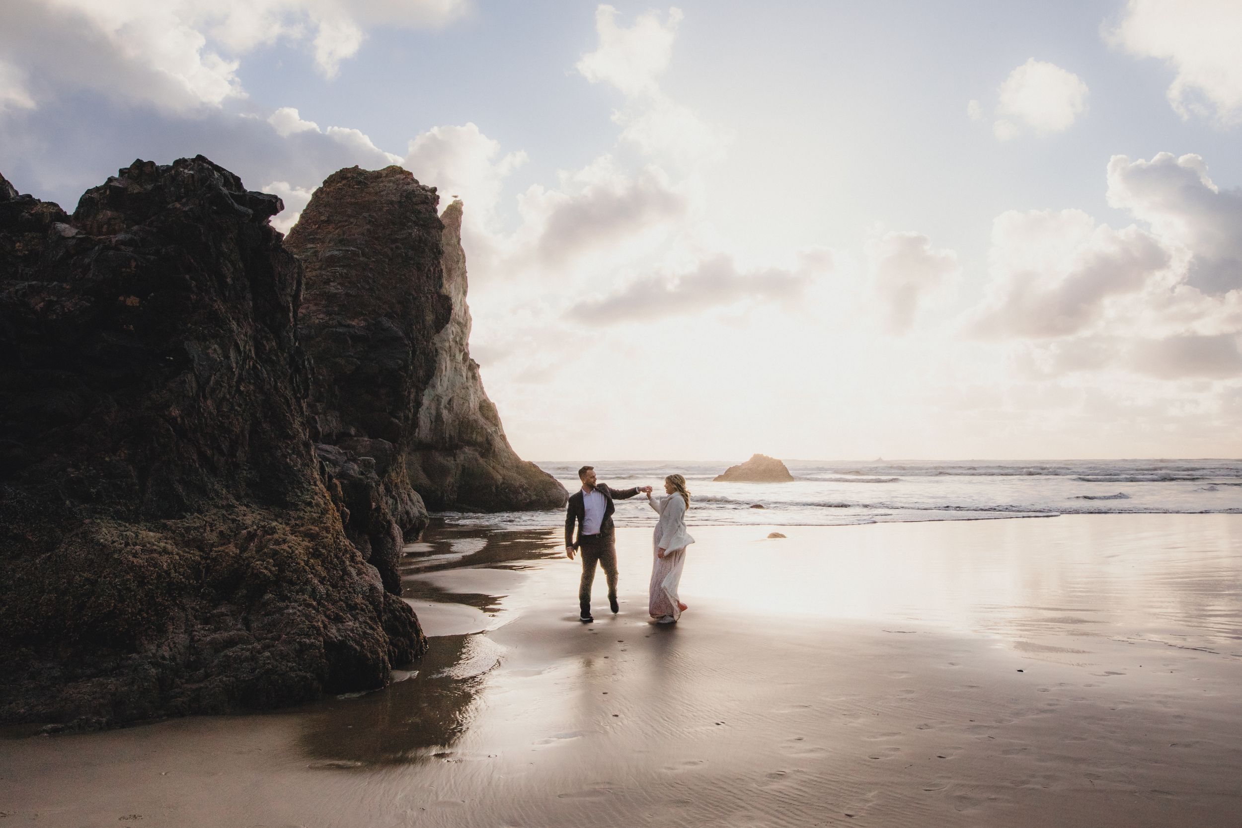 A couple walks along a scenic beach at sunset with towering cliffs in the background.