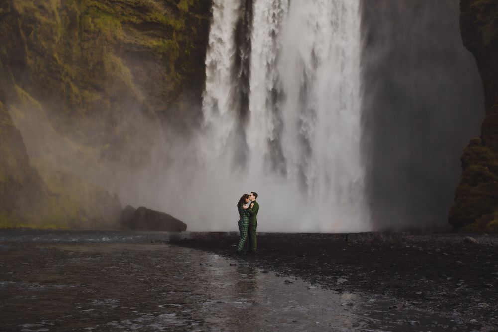 Couple embracing in front of majestic Skogafoss waterfall in Iceland, surrounded by misty spray and dark volcanic rock.