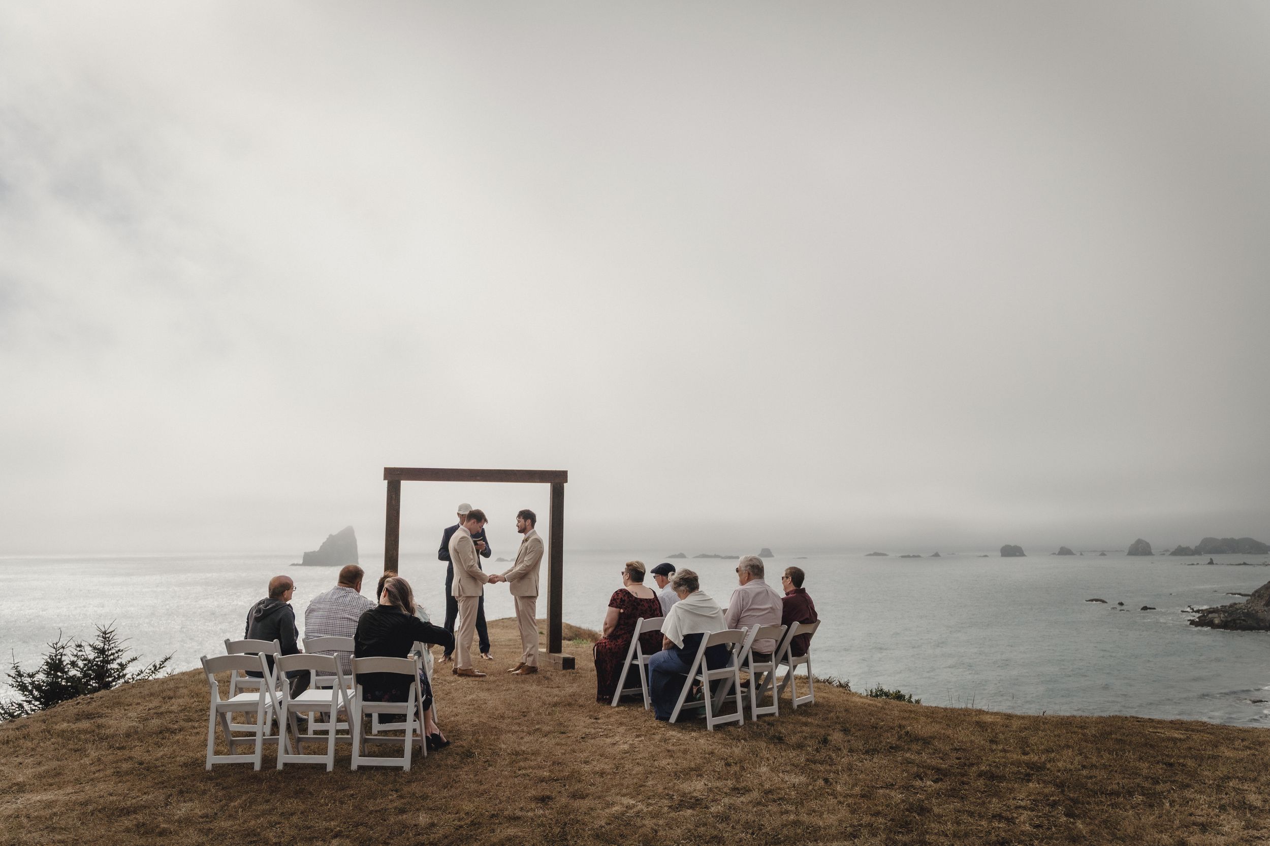 Small outdoor wedding ceremony on a coastal cliff overlooking the misty ocean with white chairs and a wooden arch.