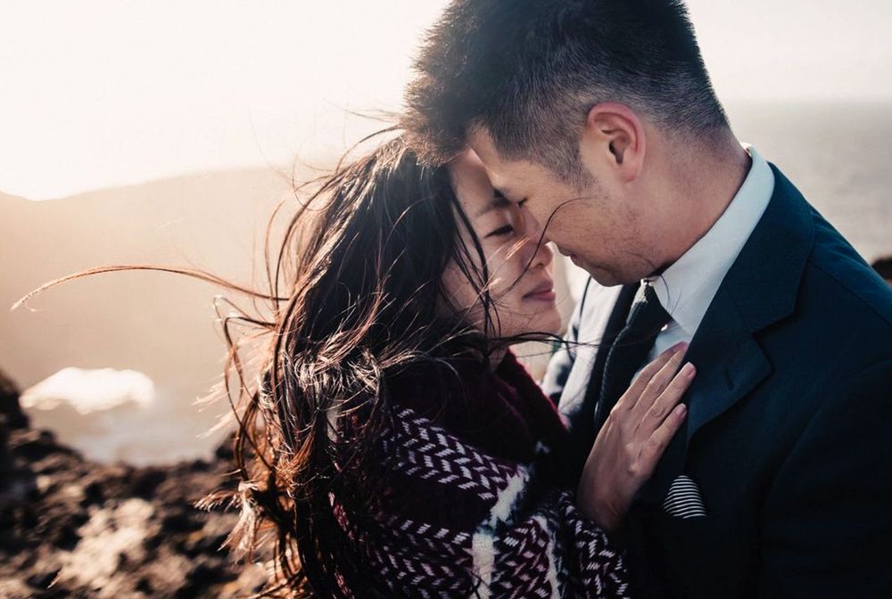 Romantic couple embracing at golden hour on a rocky hilltop, wind blowing through her hair.