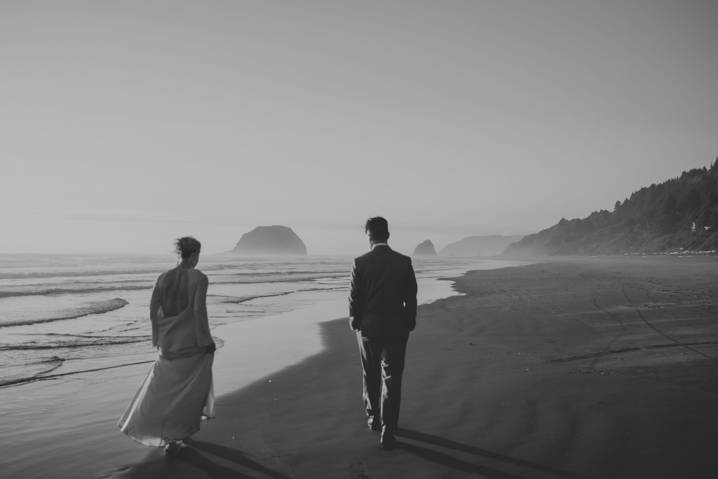 Black and white photo of a bride and groom walking along a misty beach with rocky headlands in the background.