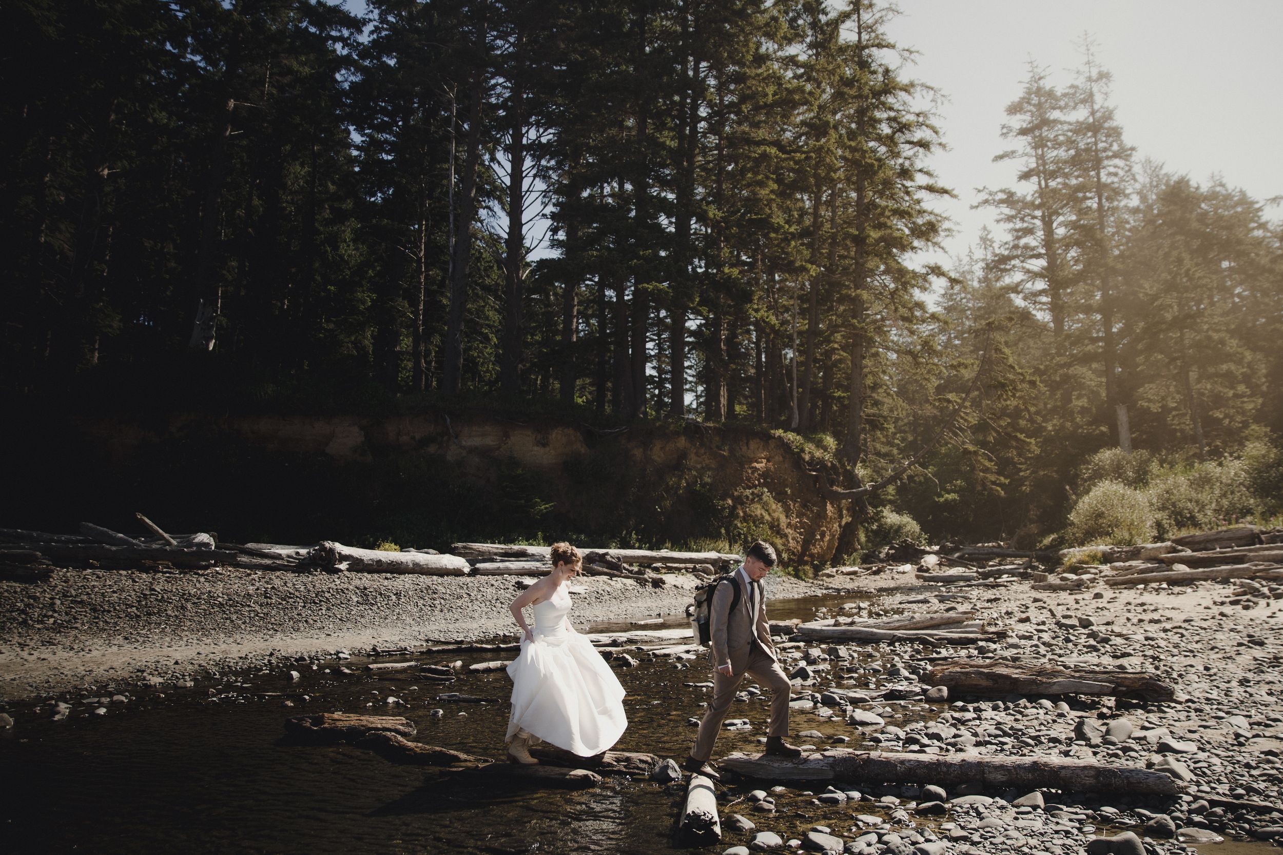 Bride and groom walk hand in hand along a rocky Pacific Northwest beach surrounded by towering evergreen trees.