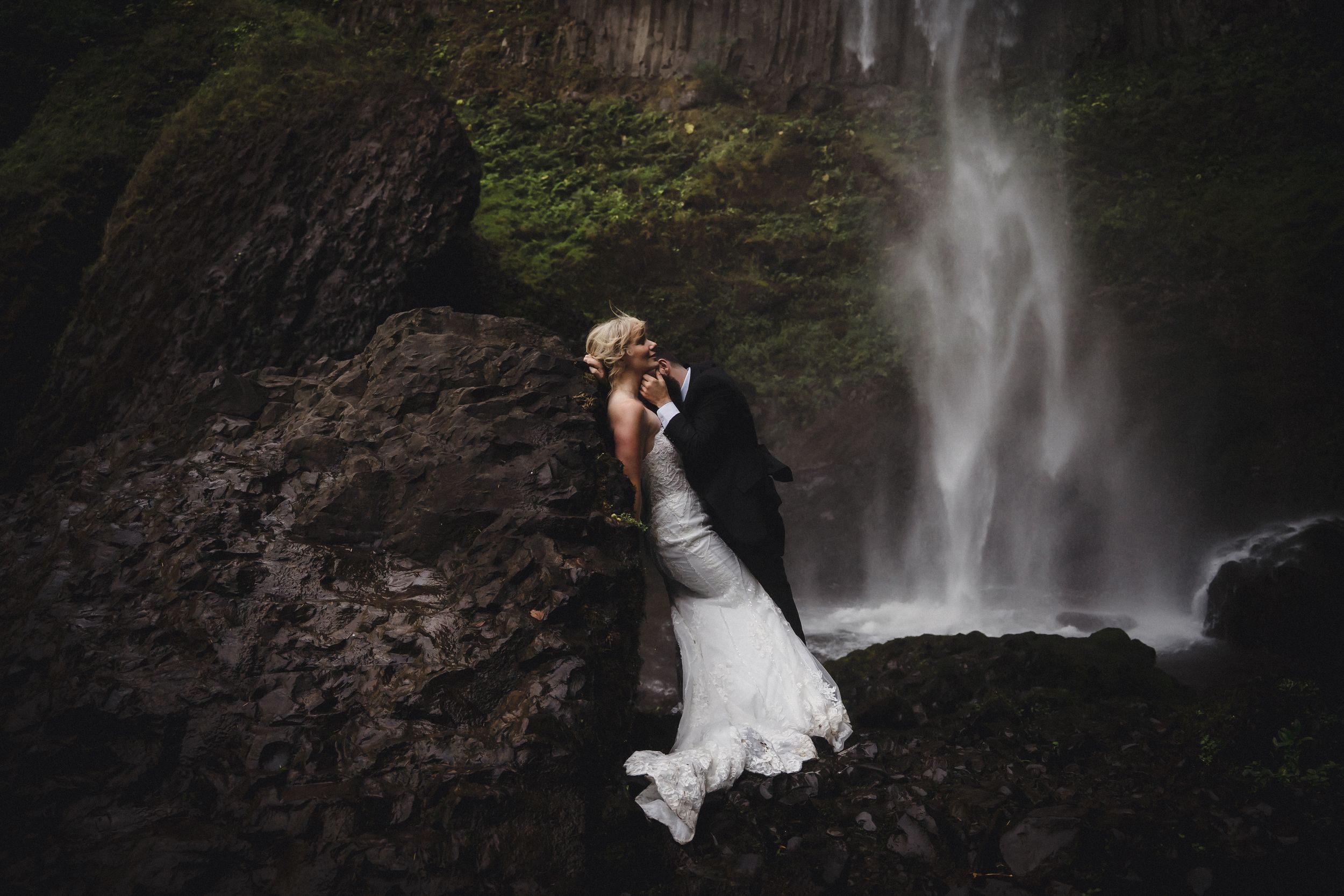 Bride and groom kissing on rocky terrain in front of a stunning waterfall, surrounded by lush greenery.