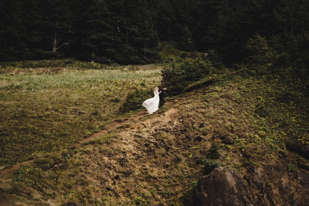 Bride in white wedding dress standing on a rocky hillside surrounded by lush green forest and golden meadow.