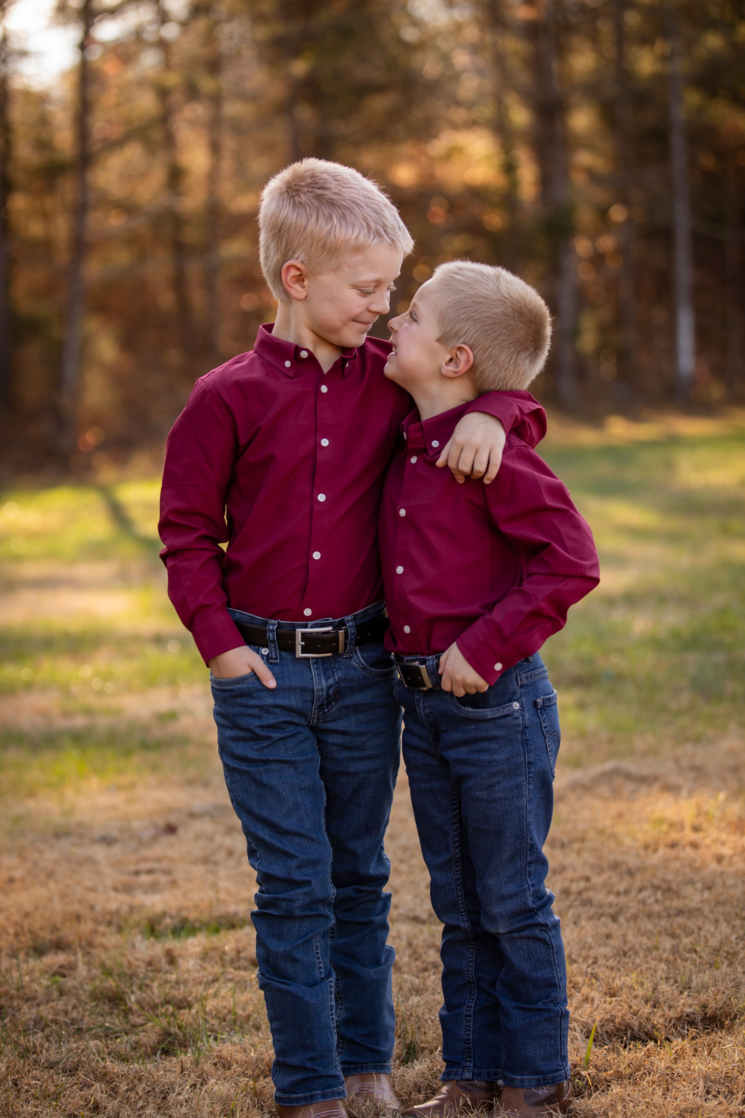 Two young children in matching burgundy shirts and jeans share a sweet moment outdoors during golden hour.