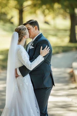A romantic wedding couple shares tender kisses in a sunlit outdoor setting with trees in the background.