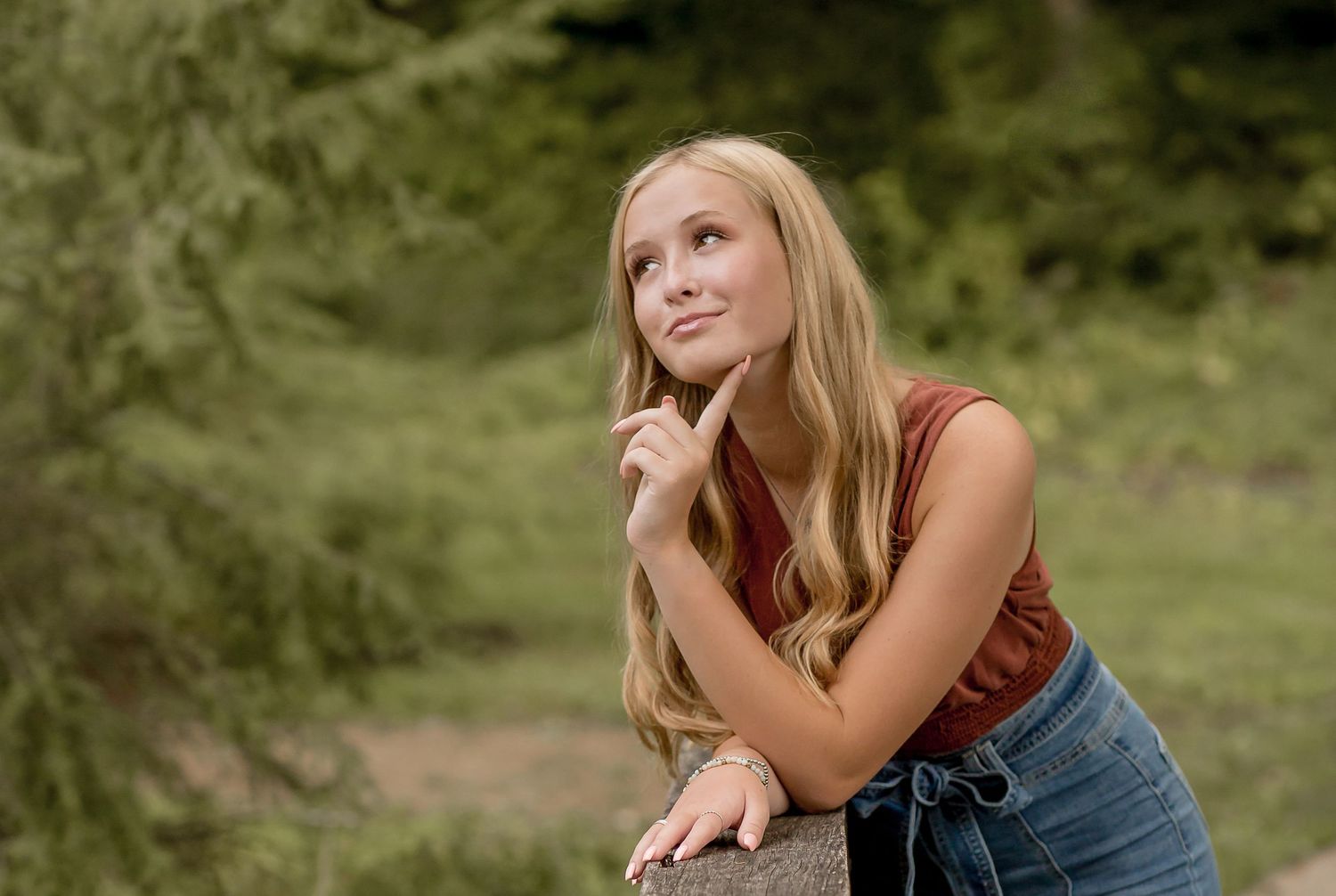 Blonde teenage girl in rust top leaning on fence gazing upward in lush green forest.