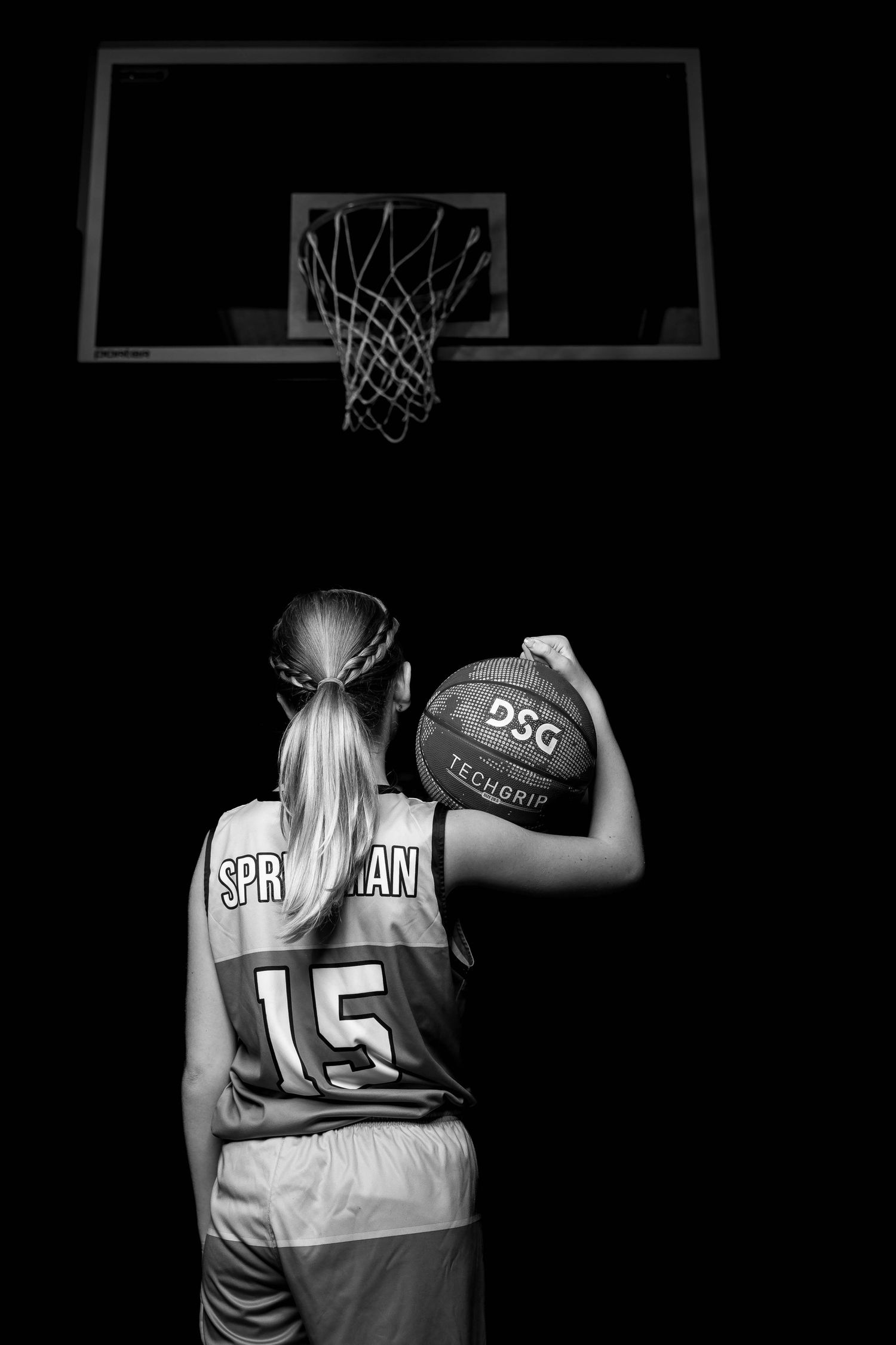 Basketball player holds ball while facing hoop in dramatic black and white composition.