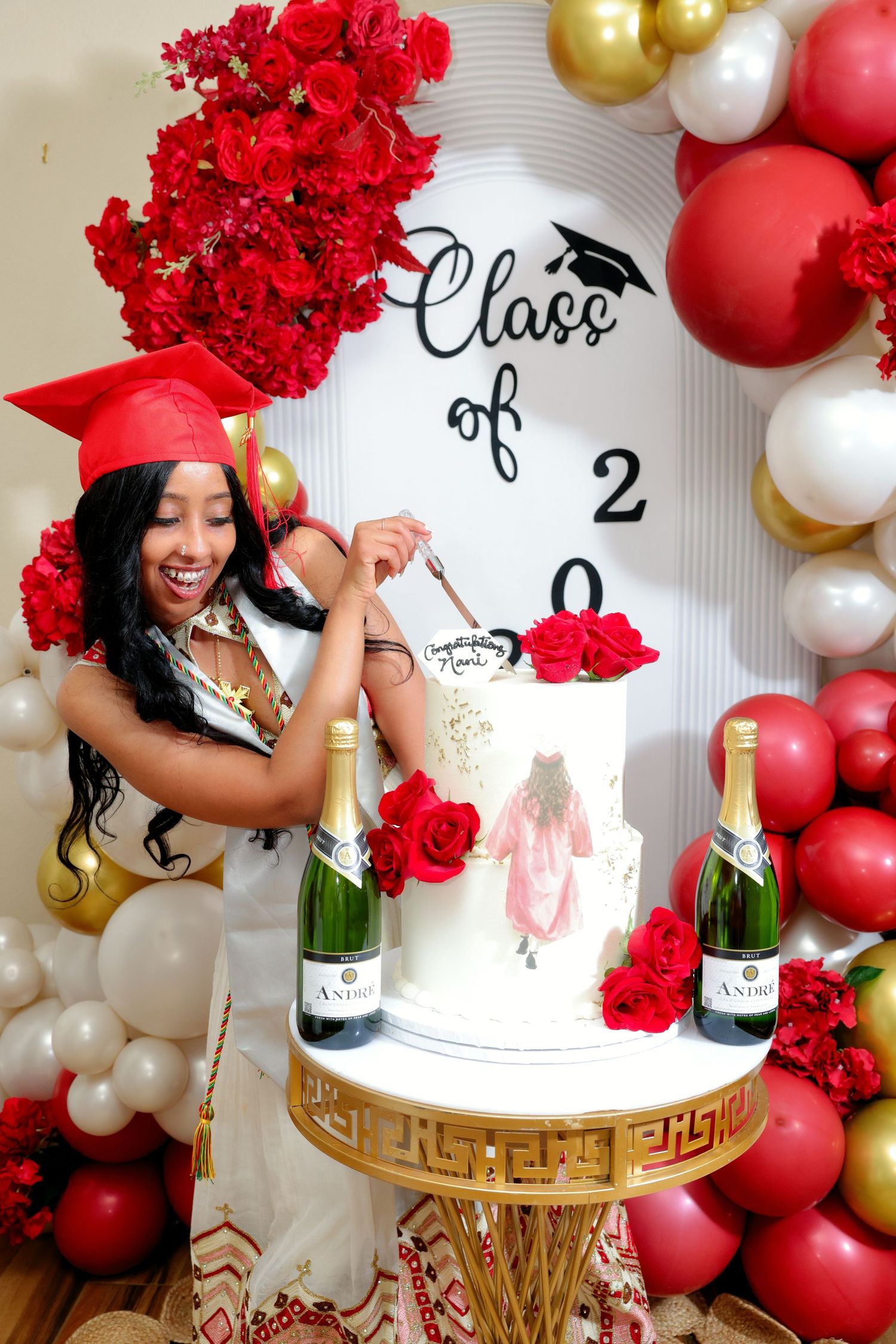 Graduate in red cap and gown cutting a decorated Class of 2026 cake with champagne bottles.