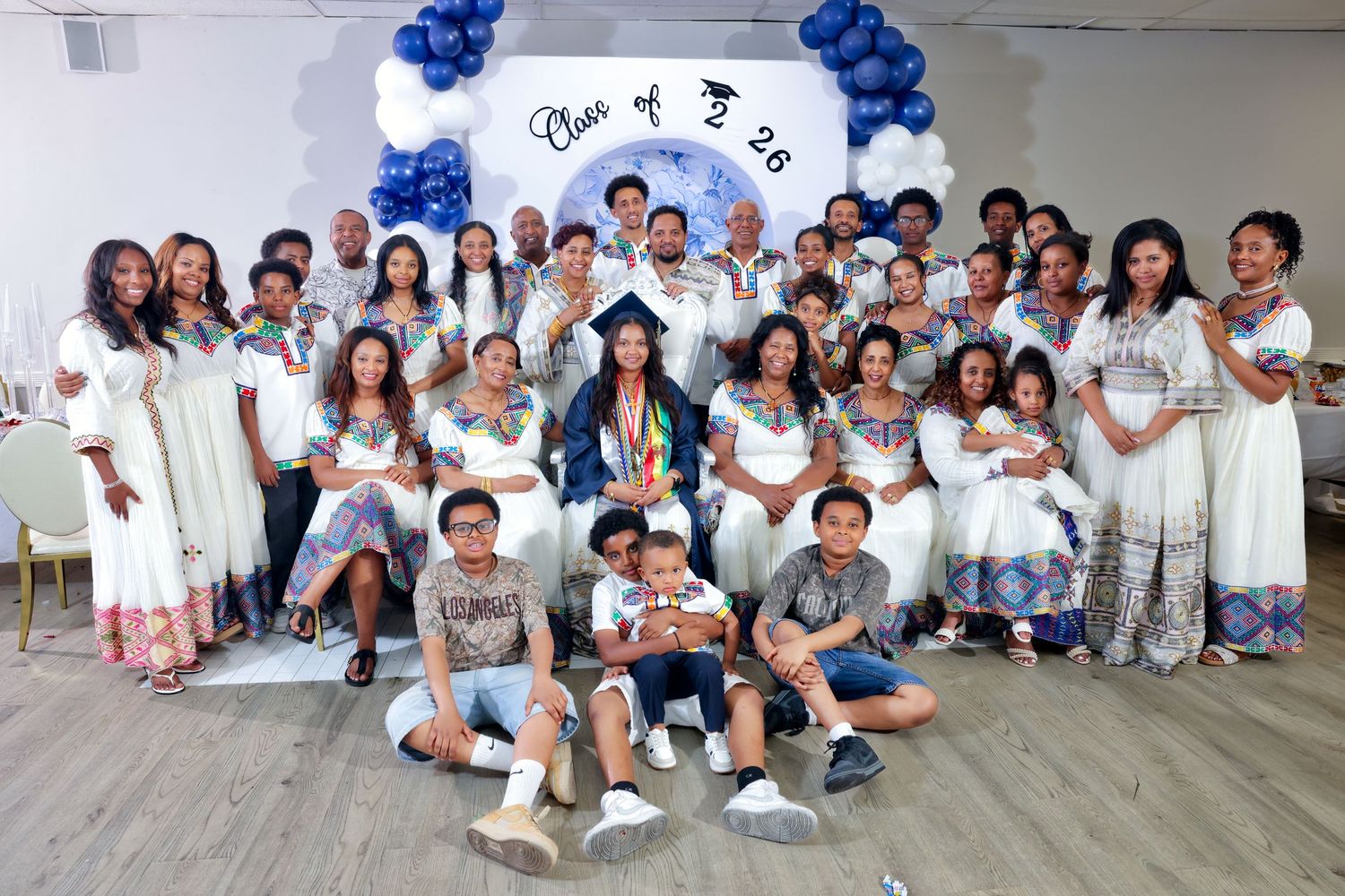Large group photo at a Class of 2026 graduation party with guests in white traditional attire.