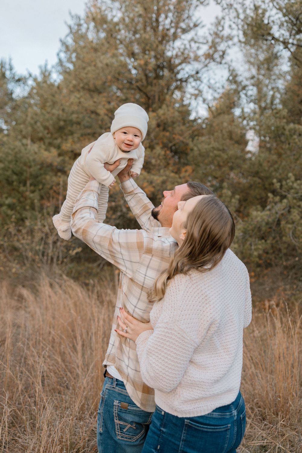 A joyful outdoor moment between parent and baby against an autumn backdrop of trees and tall grass.