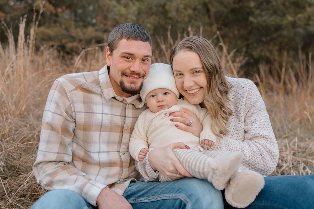A happy family of three poses together outdoors in casual attire against a natural fall setting.