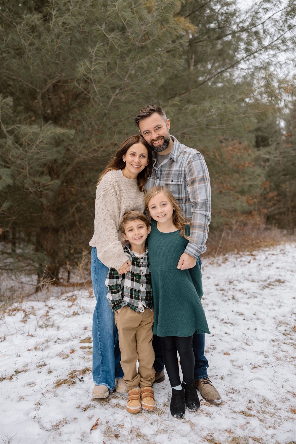 A family of four poses together on a snowy hillside with evergreen trees in the background.