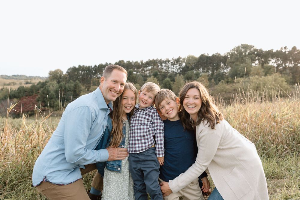 A family poses together outdoors in a field with tall grass and trees during golden hour.