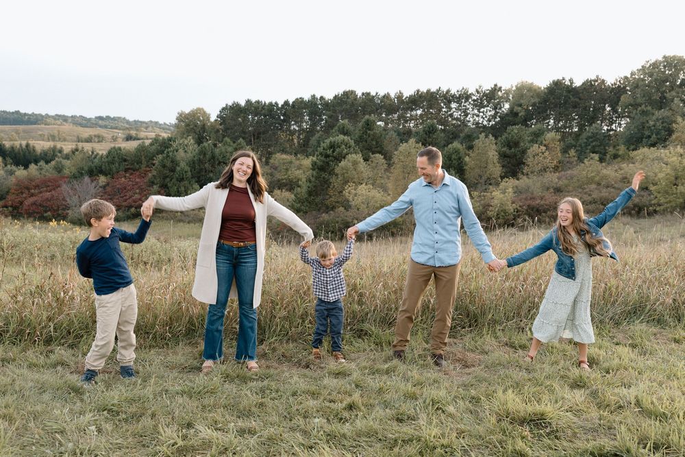 A family holds hands in a line while standing in a grassy field with trees in the background during autumn.