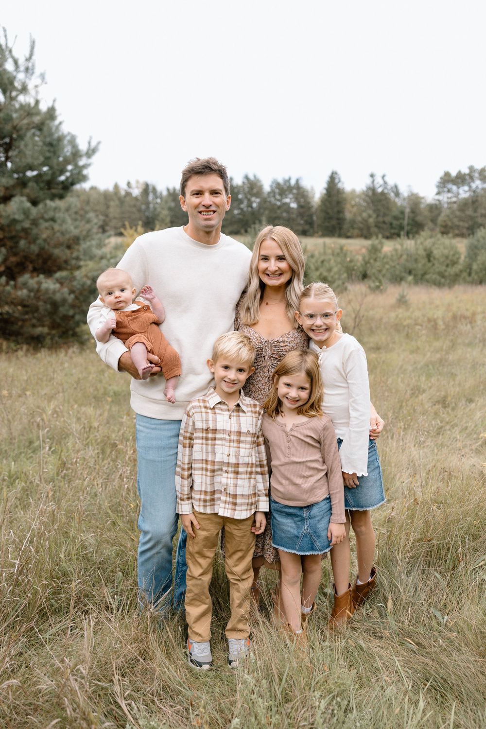 A family poses together in a natural field setting wearing coordinated casual outfits in neutral tones.