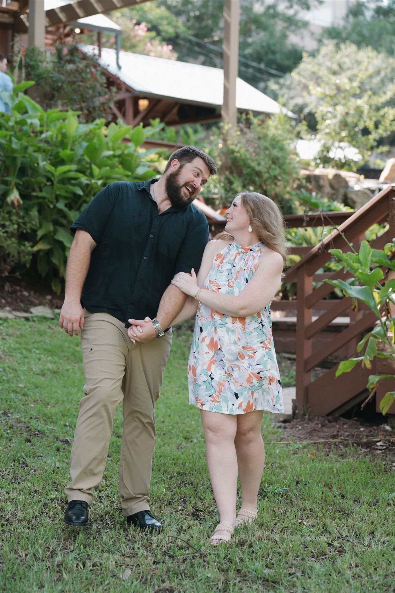 A couple shares a joyful moment together outdoors on a grassy lawn with wooden steps and greenery in the background.