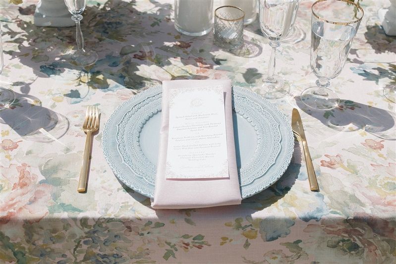 An elegant place setting with a blue charger plate, gold cutlery, and menu card on a floral tablecloth.