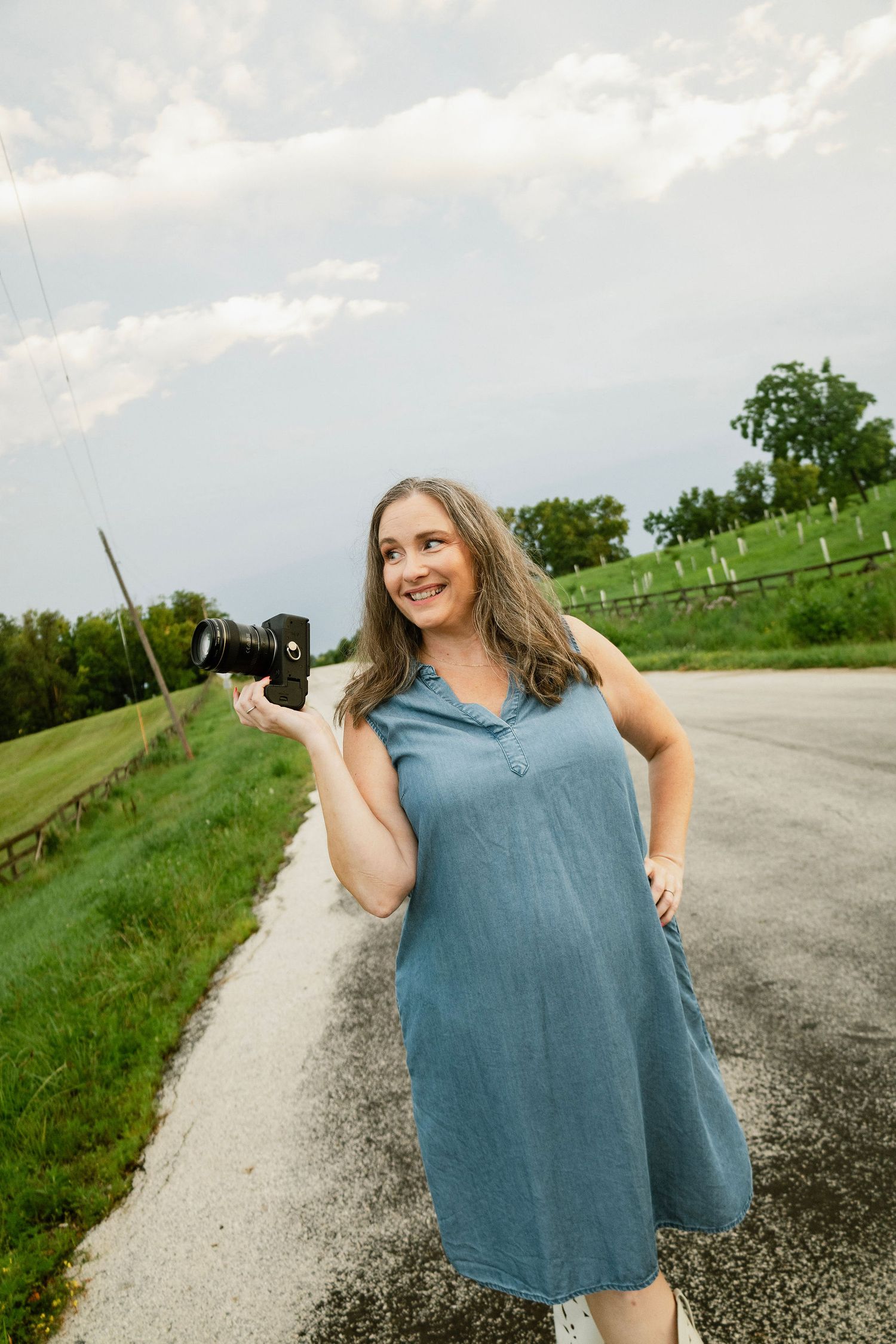 Eastern Iowa family and senior photographer Nichole Sieck Photography stands on a paved road with her camera looking toward a grassy field.