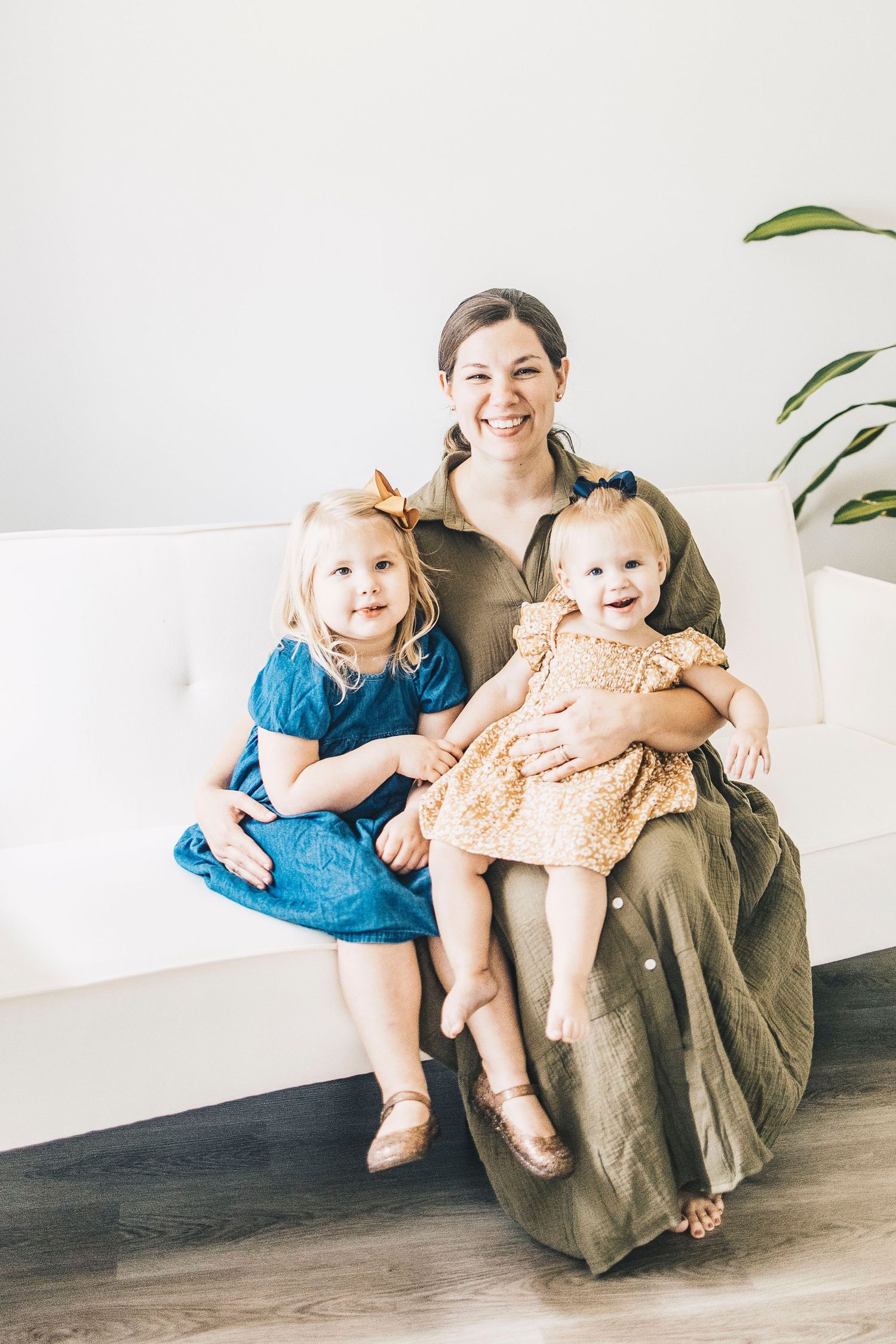 A smiling mother in olive dress sits on white sofa holding two young daughters in blue and gold dresses.