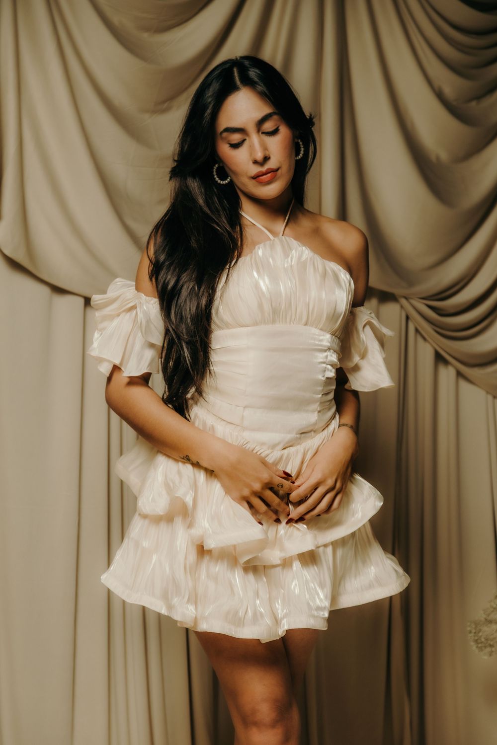 Woman in ivory shimmer off-shoulder ruffle mini dress with hoop earrings posing against beige draped curtains.