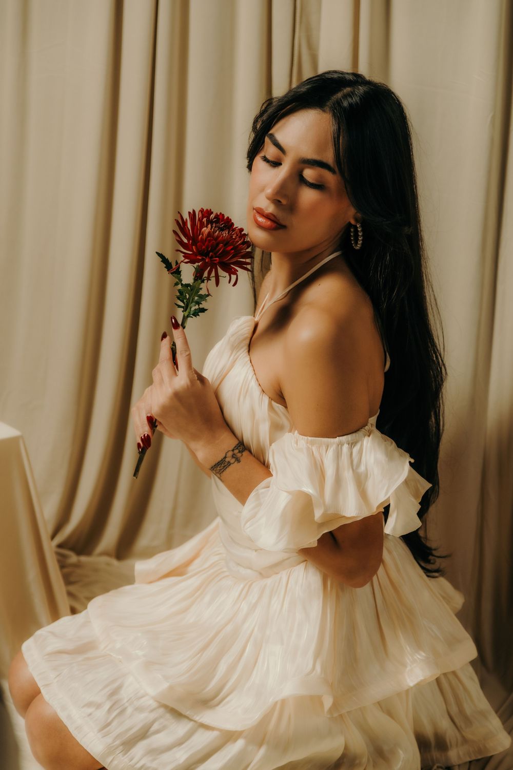 Woman in white off-shoulder ruffle dress holds red chrysanthemum flower against beige curtains.