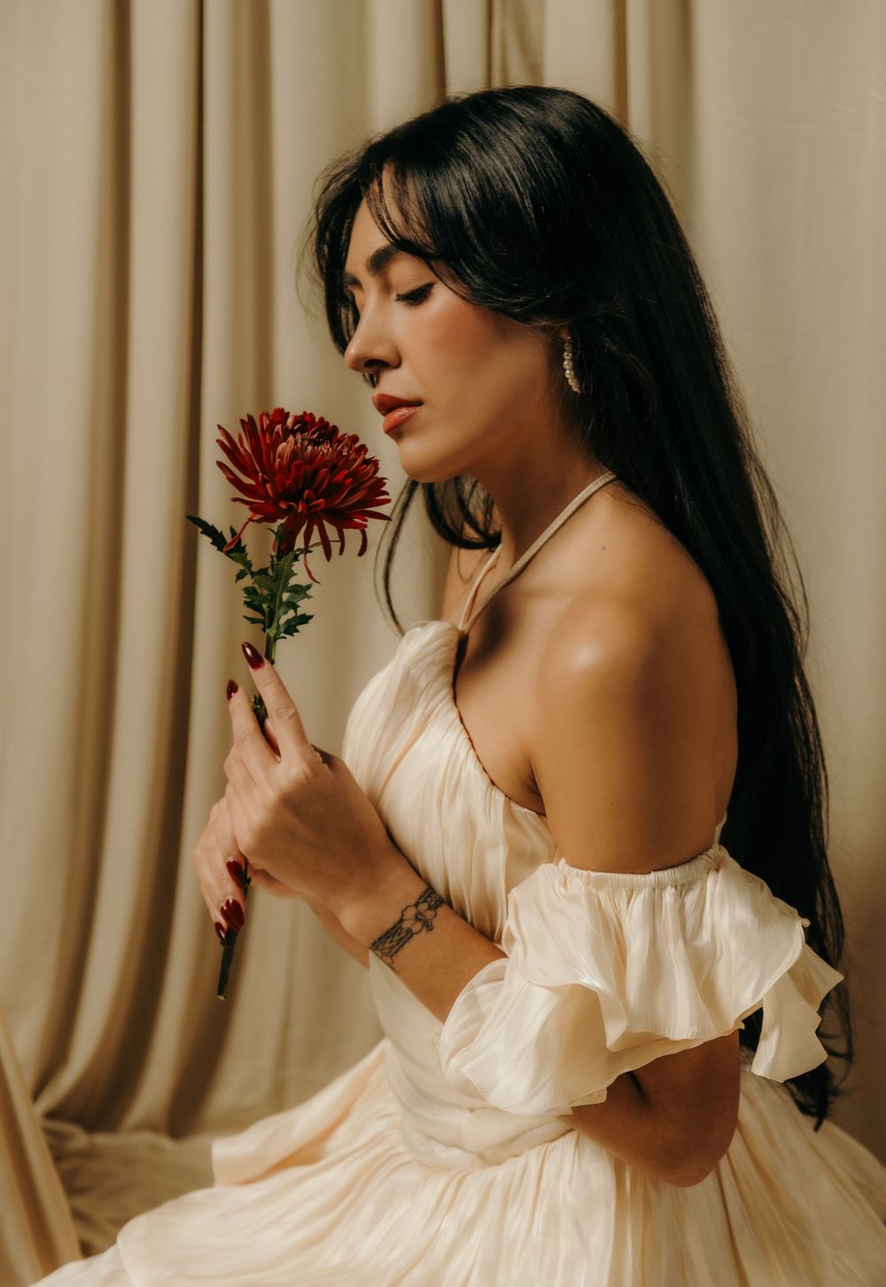 A woman in a cream off-shoulder dress holds a red chrysanthemum flower, eyes closed, posed against beige curtains.