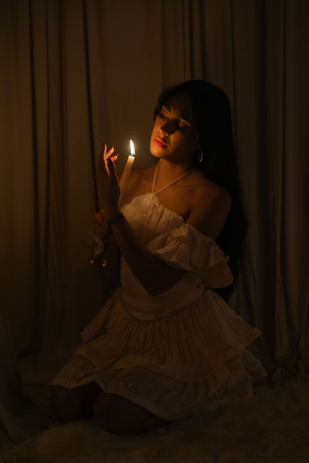 Woman in white ruffled mini dress holds glowing candle in dark moody candlelit setting.