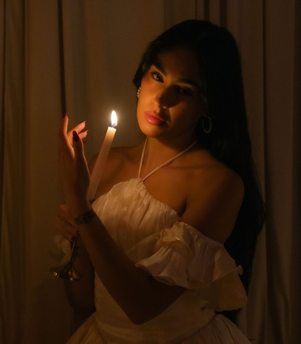 Young woman in white dress holding a lit candle in dramatic candlelight photography.