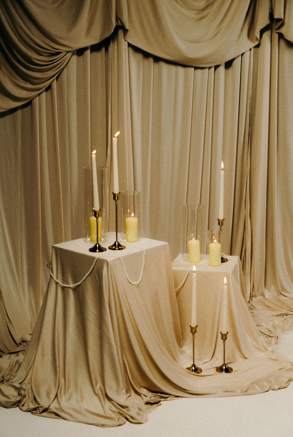 Elegant gold candlesticks with ivory taper candles on draped beige tables against luxurious flowing satin curtain backdrop.