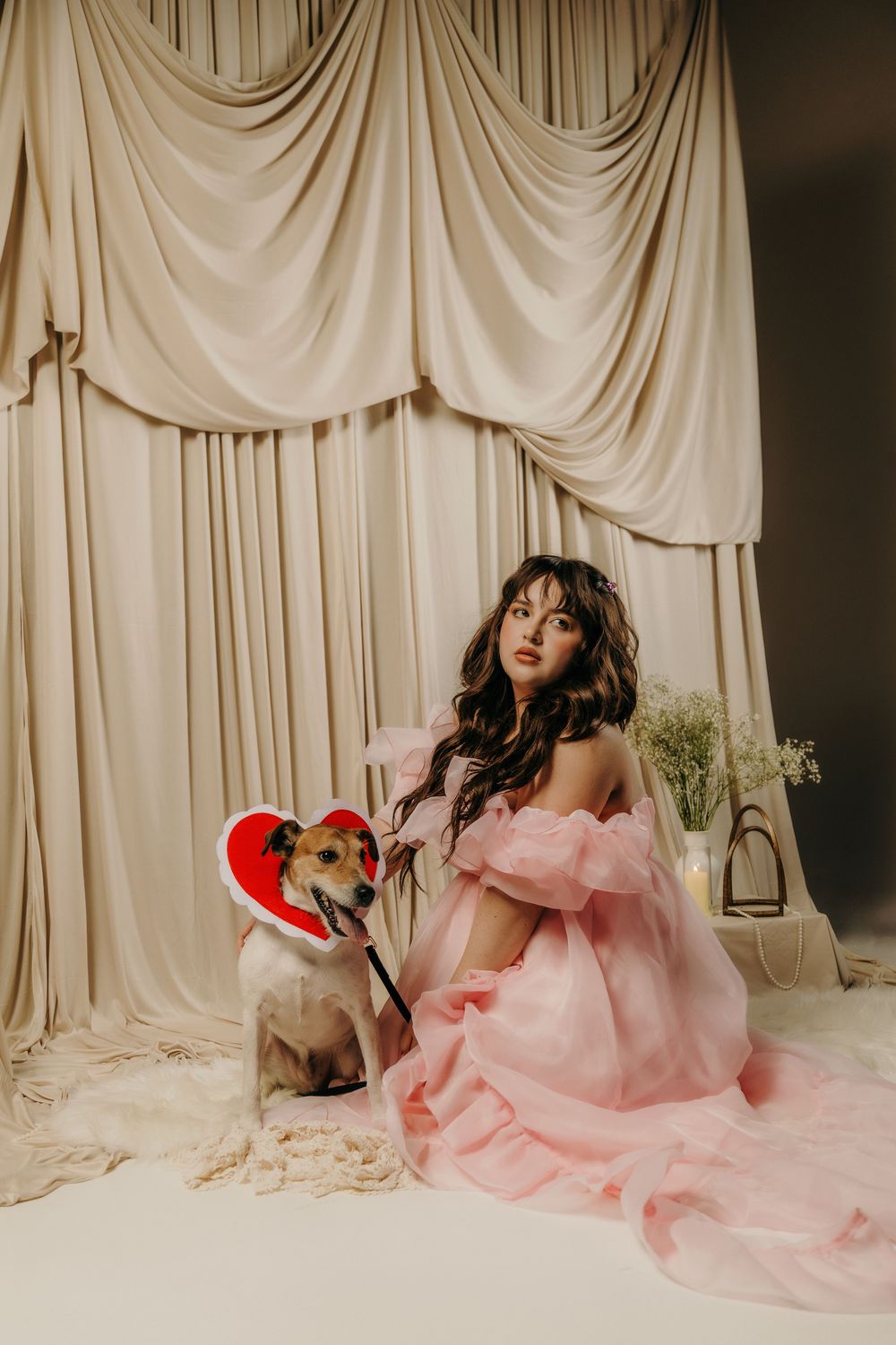 Woman in pink off-shoulder gown poses with a dog wearing a heart collar against elegant draped curtains.