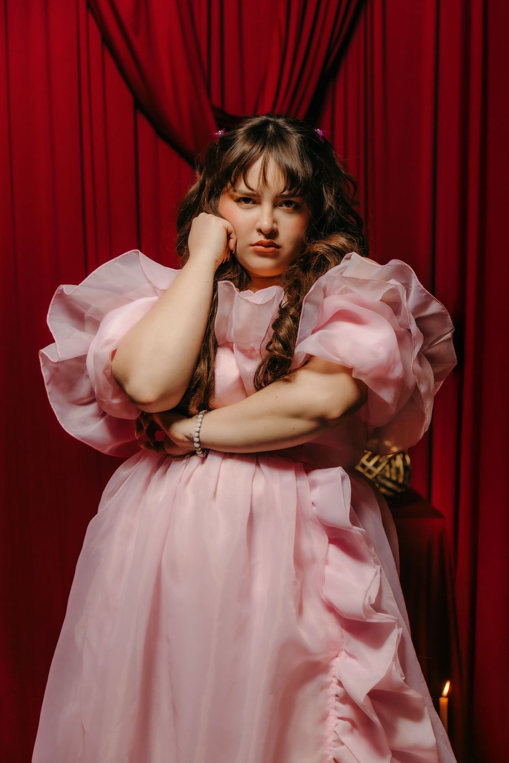 Woman in pink puffed-sleeve organza gown poses against red velvet curtains with a bored expression.