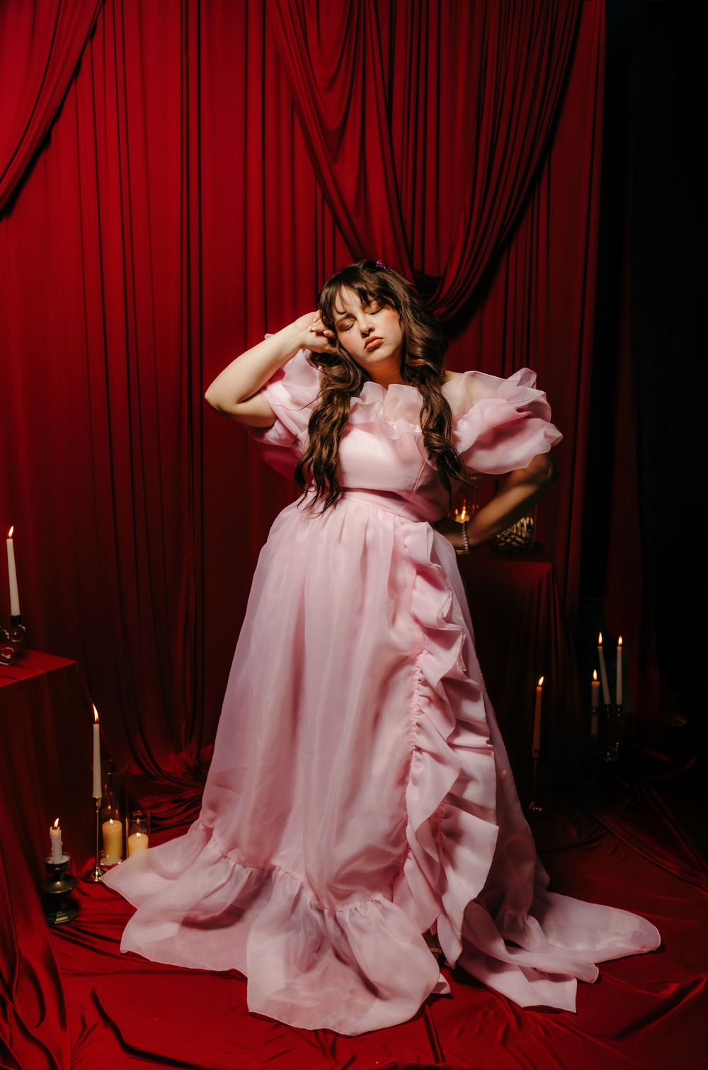 Woman in pink ruffled ball gown poses dramatically against red velvet curtains with candles.