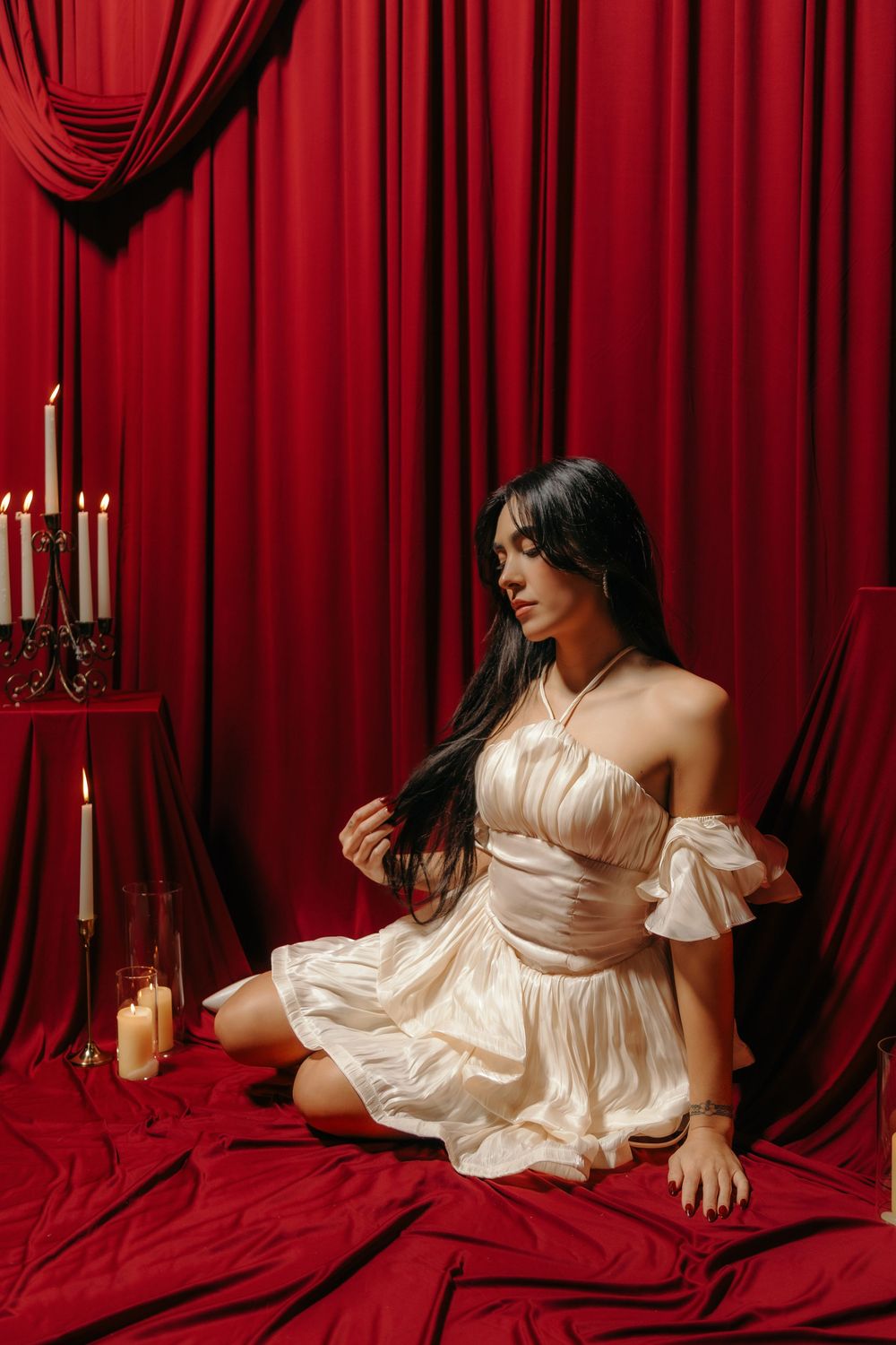 Woman in white satin dress sits on red fabric floor near candles with dramatic red curtain backdrop.