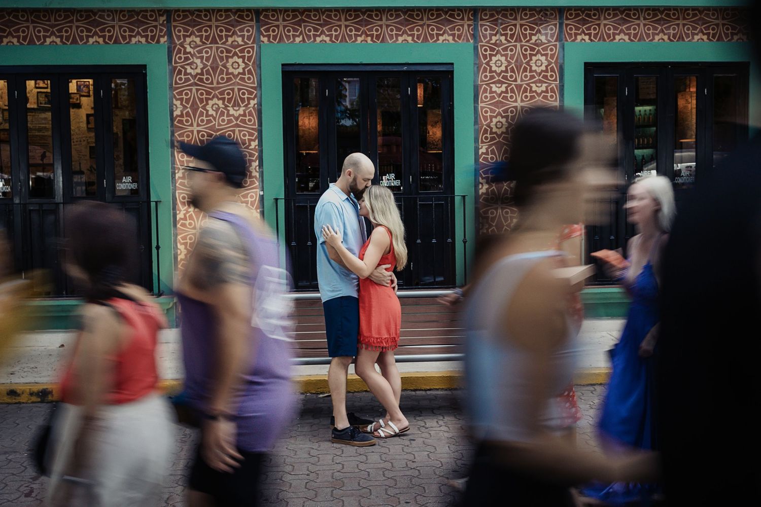 Couple embraces in front of a colorful street wall while people walk by, capturing an urban moment in Playa del Carmen.