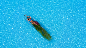 Aerial view of a person relaxing on a blue kayak with a palm leaf in a crystal clear turquoise pool.